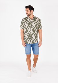 Kurzarm-Hemd mit Knopfleiste in olivgrünem und beigem geometrischem Muster, kombiniert mit Jeansshorts und weißen Turnschuhen. Glatte Stofftextur.