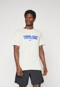 Lysebeige kortærmet t-shirt med teksten "SEJR STARTER MED TRÆNING." i blå, sammen med sorte shorts. 100% bomuldsmateriale, afslappet pasform.