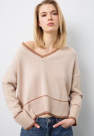 Giovane donna con capelli biondi fino alle spalle, indossa un maglione beige lavorato a maglia con rifiniture marroni e jeans blu, in piedi con le mani nelle tasche.