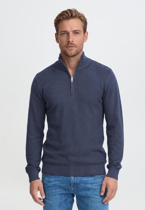 Homme portant un pull bleu marine à fermeture éclair quart et un jean bleu, debout devant un fond clair uni.