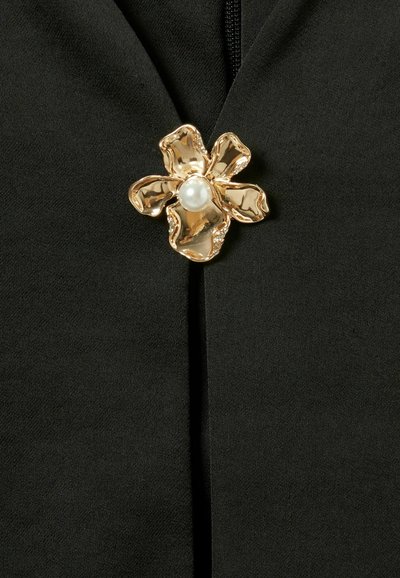 Tejido negro con un broche decorativo de flor dorada que presenta una perla central y un diseño de pétalos texturizados, realzando la apariencia de la prenda.