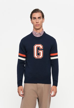 Ung mann iført marineblå genser med stor oransje og hvit "G," stripete ermer, beige bukser og en rød og hvit stripete skjorte med krage.