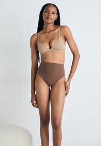 Beige myk stoff bh med tynne stropper, kombinert med høyt liv brune truser. Glatt tekstur og moderne snitt, båret av en modell.