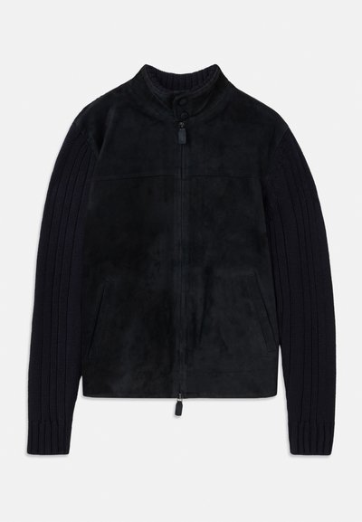 Jachetă neagră cu un corp din suede neted și mâneci tricotate texturate. Are un guler înalt, închidere cu fermoar și buzunare laterale.