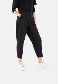 Black tailored trousers with pleats, featuring side pockets and a cropped ankle length, paired with white sneakers. Smooth texture.
