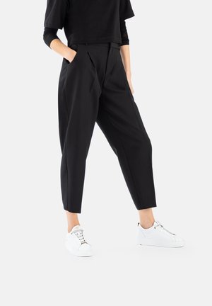 Black tailored trousers with pleats, featuring side pockets and a cropped ankle length, paired with white sneakers. Smooth texture.