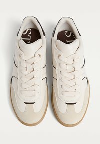 Une paire de baskets crème avec des capuchons en suède beige, des lacets blancs, des détails en noir et des semelles en caoutchouc marron clair sur un fond blanc.