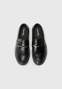 Mocasini negri din piele cu un finisaj neted, o bandă largă peste partea superioară a piciorului și un detaliu metalic. Numele brandului este vizibil pe tălpic.
