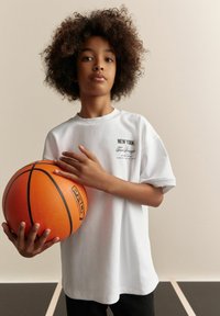 Hvid t-shirt med korte ærmer og teksten "NEW YORK" på brystet, båret af et barn, der holder en orange basketball med tekstureret overflade og sorte linjer.