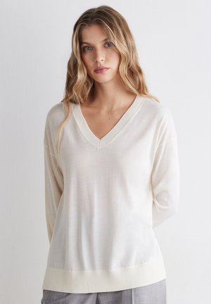 Vrouw met lang blond haar die een witte trui met V-hals en grijze broek draagt, staat voor een effen witte achtergrond.