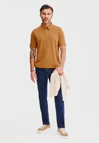 Brązowa koszulka polo, granatowe dżinsy, jasne buty, z kremowym swetrem zarzuconym na ramię; swobodny krój, krótkie rękawy, jednolity kolor.