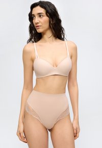 Γυναίκα φοράει σουτιέν σε γυμνό χρώμα με λεπτές τιράντες και σλιπ ψηλόμεσο με δαντέλα, στέκεται μπροστά σε απλό φωτεινό φόντο.