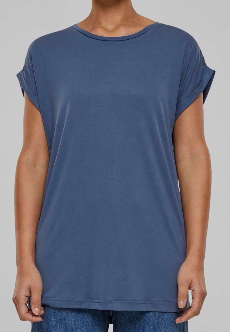 Personne portant un t-shirt bleu à manches courtes ample et un jean bleu, debout devant un fond gris uni.