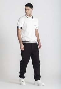 Camisa polo blanca con diseño de punto texturizado, cuello negro y franjas en las mangas, combinada con pantalones negros y zapatillas blancas.