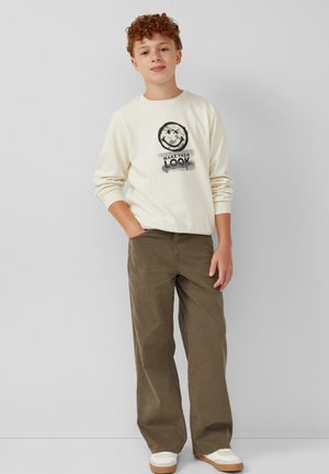 Garçon aux cheveux roux portant un sweat-shirt crème avec un smiley et du texte, un pantalon large olive et des baskets blanches, se tenant contre un mur uni.