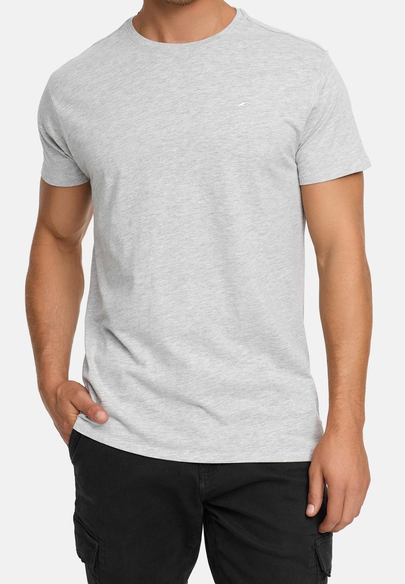 Homme portant un t-shirt gris clair uni à manches courtes et un pantalon noir avec poches, debout avec une main dans la poche.