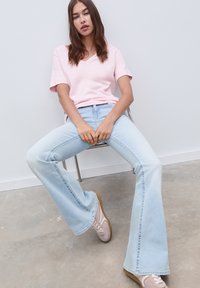 Helles rosa V-Ausschnitt-T-Shirt, kombiniert mit hellblauen, ausladenden Jeans, die subtile Verblassungen aufweisen. Das Model sitzt auf einem Stuhl und trägt hellfarbene Sneakers.
