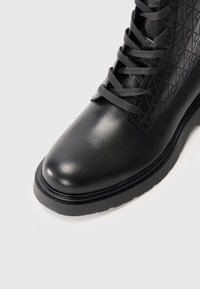 Botín negro con cordones hasta el tobillo, con una superficie de cuero liso y un panel con textura de relieve. Presenta una puntera redonda y una suela de goma duradera.