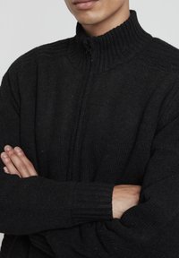 Pulover negru din tricot, cu guler înalt și manșete cu textură. Se închide cu fermoar și are un model texturat pe umeri. Material moale.