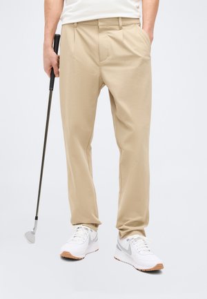Homme portant un pantalon beige et des baskets blanches, tenant un club de golf d'une main et ayant l'autre main dans la poche, sur un fond uni.