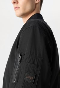 Černá bomber bunda vyrobená z lehké tkaniny, s kapsou na zip na rukávu a logem. Hladká textura a přiléhavý střih.