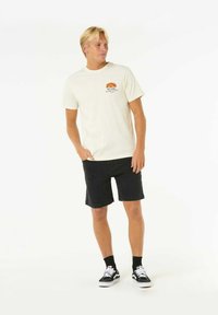 Cremefarbenes Baumwoll-T-Shirt mit einem bunten Retro-Sonnenuntergangs-Graphic, kombiniert mit schwarzen Jeansshorts und schwarz-weißen Sneakers.