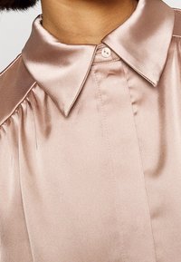 Blouse en satin rose pâle avec un col pointu classique, fermeture boutonnée sur le devant et détails froncés aux épaules. Texture lisse et brillant.