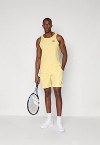 Helles gelbes Tanktop und Shorts aus weichem Material, mit einem Logo oben links. Das Model hält einen Tennisschläger und trägt weiße Turnschuhe.