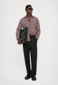 Schwarze Herren-Hosen, kariertes bordeauxfarbenes Hemd, hält eine schwarze Lederhandtasche, trägt schwarze Sonnenbrillen, kombiniert mit schwarzen Sneakers.