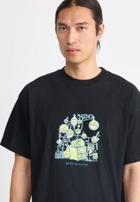 Sort bomulds t-shirt med et farverigt grafik af videnskabsrelaterede illustrationer, herunder kolber og smilende figurer, med teksten "BEDST I KLASSEN".