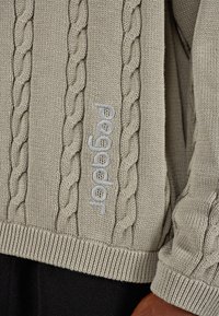 Svetlo sivi pleteni pulover z vzorcem kabla in rebrastim robom. Na strani je izvezena blagovna znamka "Pegador" v usklajeni barvi. Mehka tekstura.