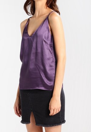 Femme portant un haut en satin violet sans manches et une jupe en jean noire avec une fente devant et un ourlet effiloché sur fond blanc.