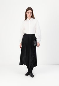 Balta pogām augšā krekls ar garām piedurknēm, kombinēts ar melnu plisētu midi svārku. Melna rokassoma un tumšas kurpes. vienkāršs, elegants dizains.