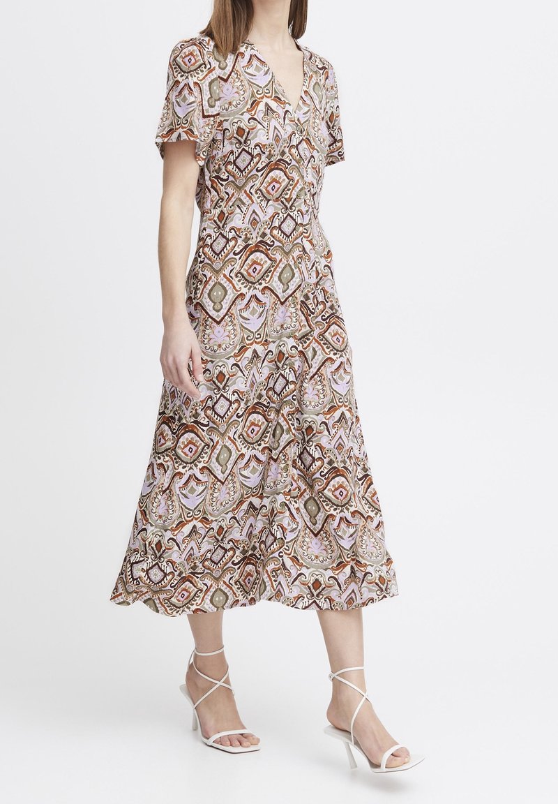 Robe maxi à col en V, manches courtes, avec un motif géométrique coloré en nuances de rose, marron et vert. Associée à des talons blancs à lanières.