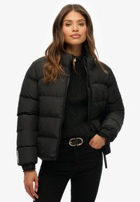 Zwarte pufferjack met hoge kraag, ritssluiting en elastische manchetten. Textuurstof over een aangesloten silhouet, gecombineerd met een zwarte trui en riem.