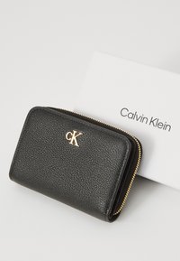 Portefeuille en cuir noir texturé avec zip doré et logo. Forme rectangulaire compacte, accompagné d'une carte blanche portant le texte "Calvin Klein".