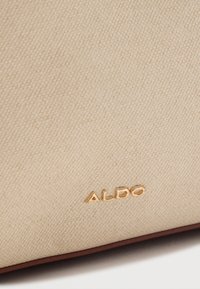 Tissu beige texturé avec de petites lettres en métal doré formant le mot "ALDO" près du bord inférieur de la surface du tissu.