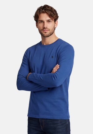 Mann mit kurzen lockigen Haaren, der ein blaues langärmeliges Hemd trägt und die Arme vor einem einfarbigen hellen Hintergrund verschränkt hat.