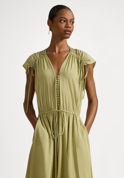 Γυναίκα με ελαφρύ ελαιδί φόρεμα, κοντά σούρα μανίκια, κουμπωτό μπροστινό μέρος, ζώνη με κορδόνι στη μέση και χέρια στις τσέπες, κοιτάζοντας προς τα δεξιά.