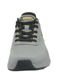 Scarpa da ginnastica grigia con tomaia in mesh testurizzata, lacci neri e accenti gialli. Caratterizzata da un design elegante e una suola in gomma con una leggera curvatura.