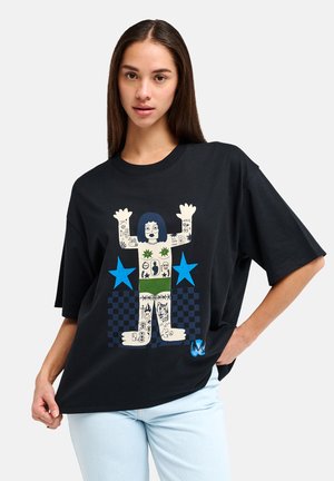 Mladá žena v čiernom oversized tričku s farebným tetovaným motívom, modrými hviezdami a vzorom šachovnice, kombinovanom s svetlomodrými džínsami.