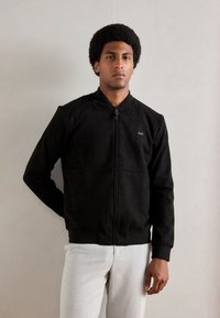 Black zip-up bomber jacket in soft fabric, featuring a high collar, ribbed cuffs, and a subtle embroidered logo on the left chest.
