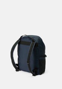 Sac à dos en nylon bleu marine avec une forme arrondie, des bretelles noires réglables et une poche latérale en mesh. Texture lisse avec des détails de couture renforcés.