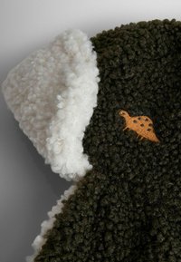 Giacca verde pelosa con colletto bianco texturizzato e ricamo di un dinosauro arancione sulla parte anteriore. Materiale morbido e peluche con un aspetto accogliente.