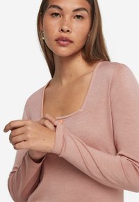 Ljusrosa långärmad tröja med rundad halsringning, ribbade ärmslut och en mjuk textur. Närbild av händer som håller i tyget.