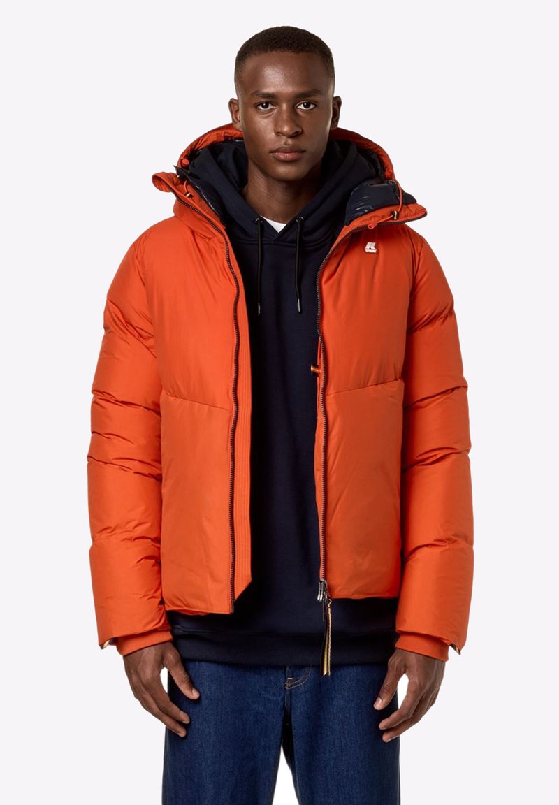 Veste matelassée orange pour homme avec capuche, dotée d'une fermeture éclair à l'avant, de deux poches et de poignets côtelés. Portée sur un sweat à capuche sombre et un jean.