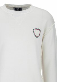 Vitt stickat tröja med ribbad rundhalssnitt och en liten sköldlogotyp på vänster bröst i grönt, rött och vitt. Texturerad tyg.