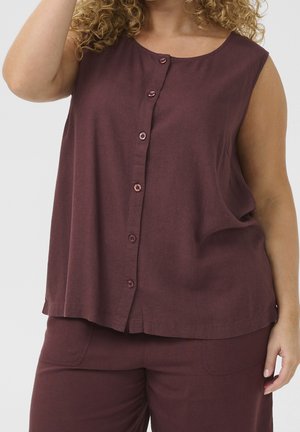 Ærmeløs knap-op top i vinrød, båret med matchende vinrøde bukser, der viser maven og en del af en krøllet persons arm og hånd.