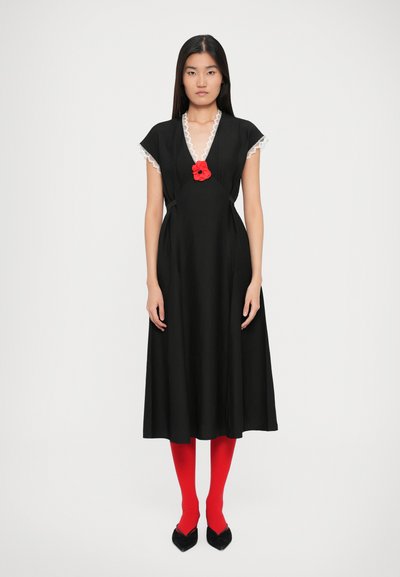 Robe noire avec des manches courtes en forme de cap, bord en dentelle et un accent de fleur rouge au niveau de l'encolure. Associée à des collants rouges et des chaussures noires à bouts pointus.