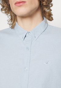 Ljusblå bomullsskjorta med knapp-down krage, matchande knappar och en broderad logotyp. Slät textur och avslappnad design.